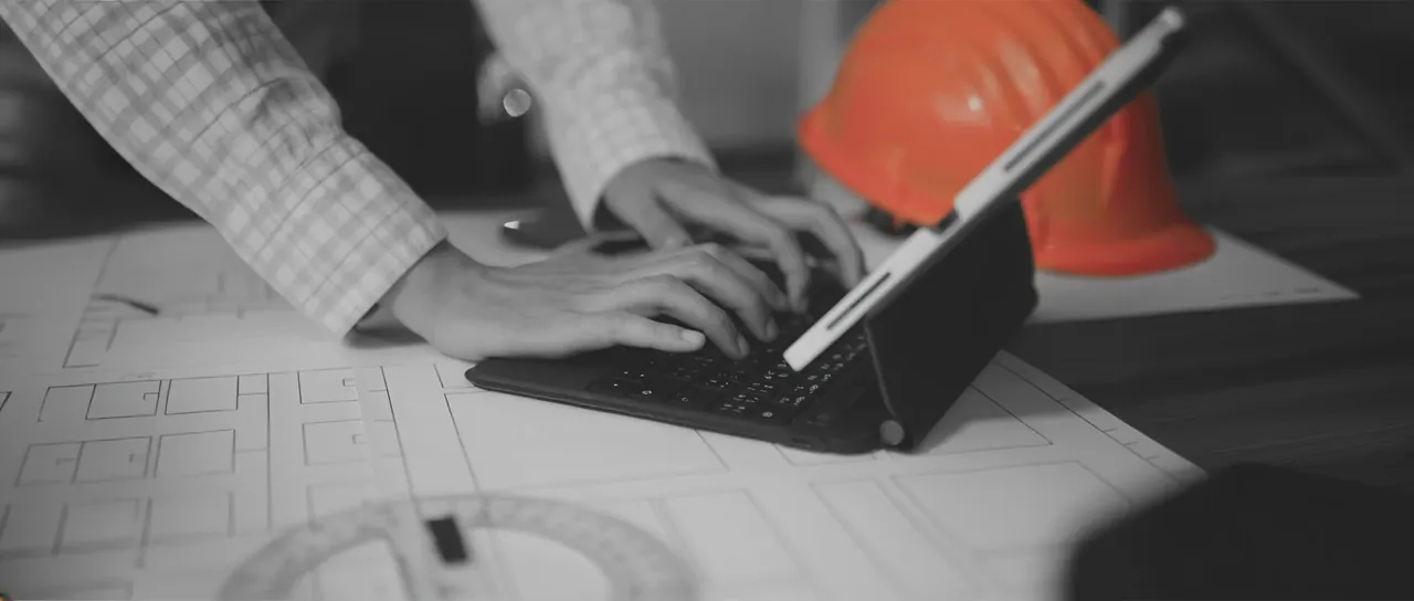 Hands typing on a tablet keyboard over construction drawings beside an orange hard hat