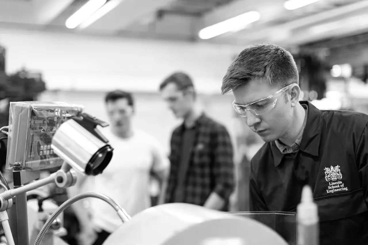 A young man in safety glasses and a dark work jacket concentrating at a lathe, with two colleagues talking in the workshop behind him