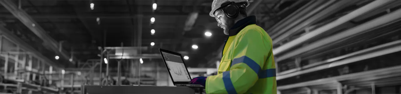 An engineer in a hard hat and ear defenders reviewing figures on a laptop in a plant hall