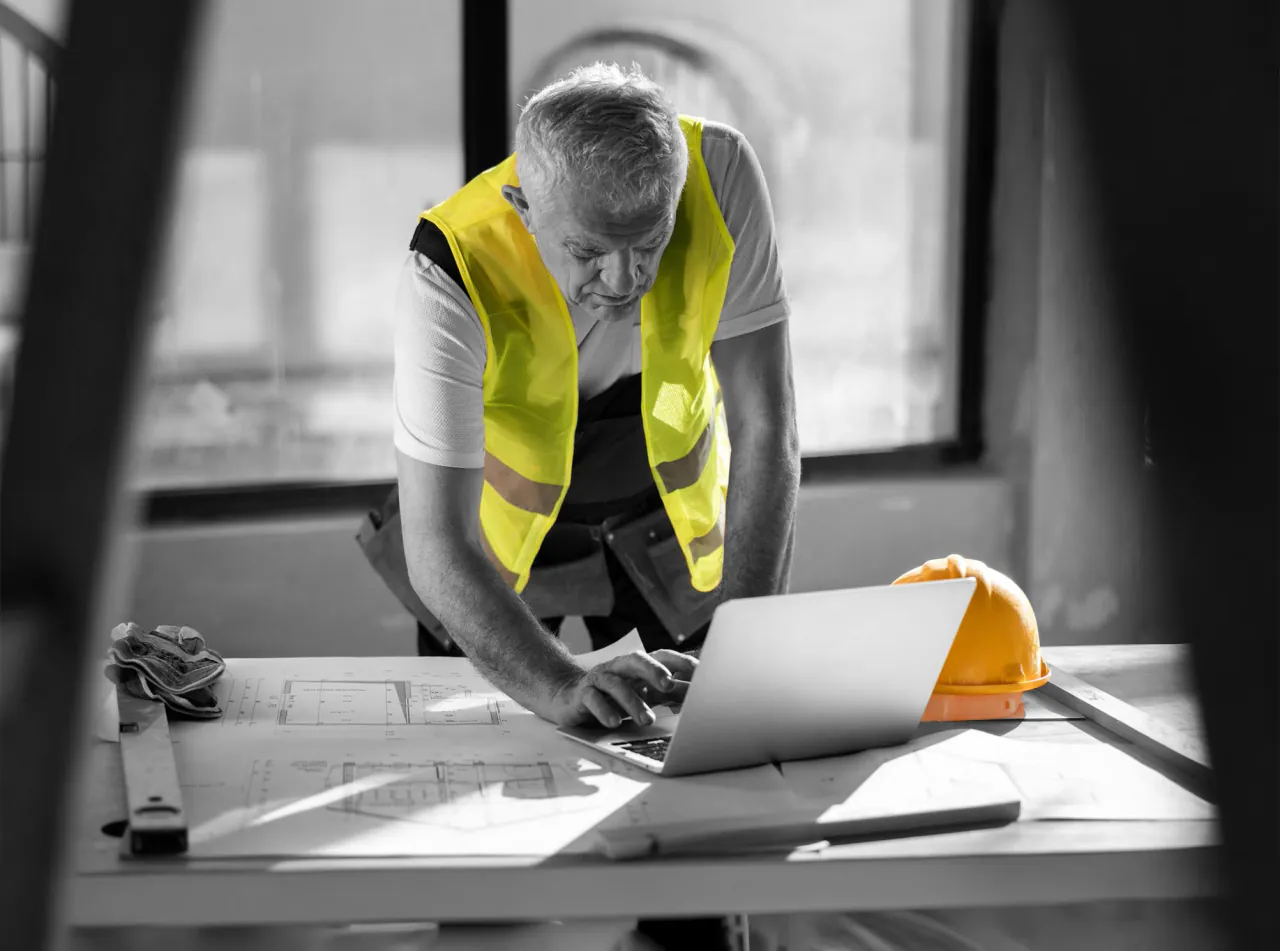 A site manager in a hi-vis vest working on a laptop over a desk of drawings