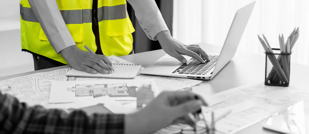 Hands typing on a laptop beside a set of drawings, a hi-vis vest in the background